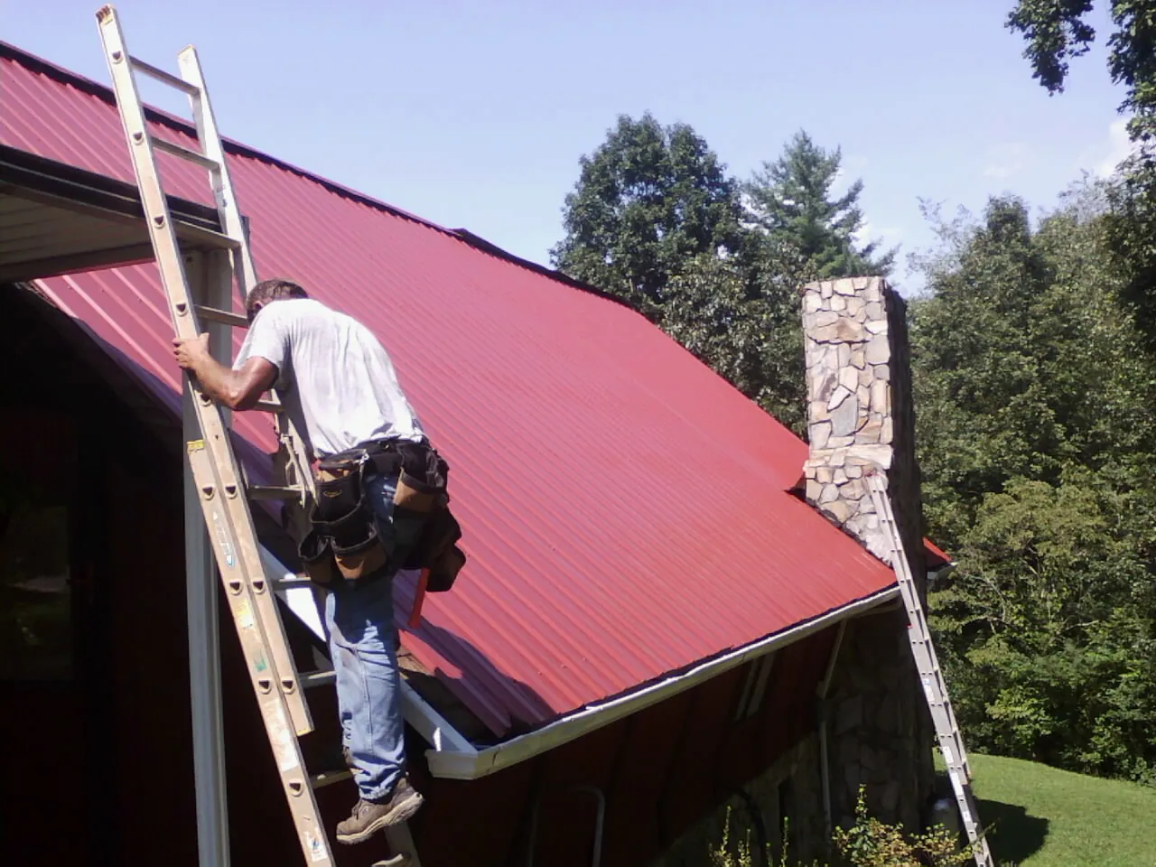 Roofer on a ladder beside a red metal roof and stone chimney — Soffit & Fascia Repair by Roofing Brothers in Mary Esther